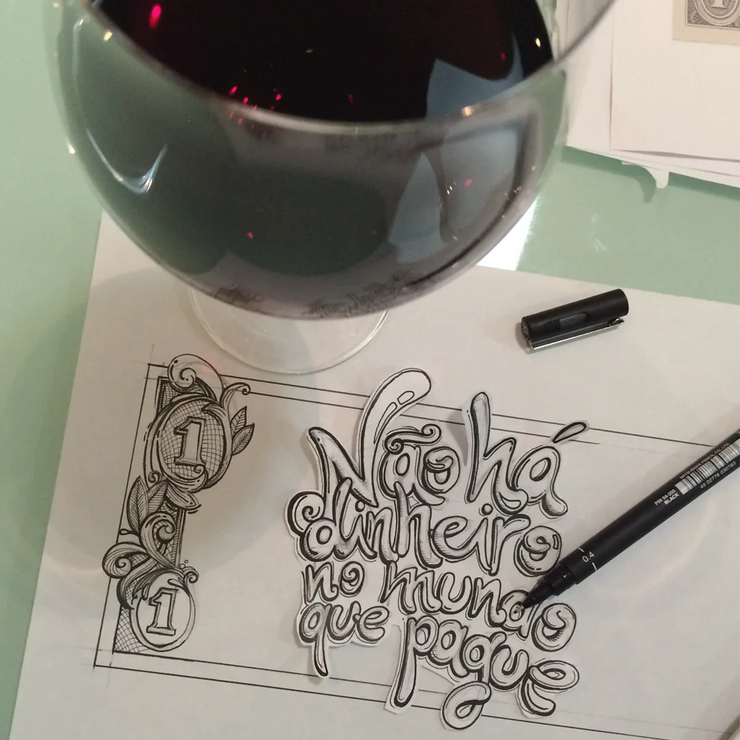 A glass of red wine beside a pen lettering sketch on paper — the manual beginning of the idea.